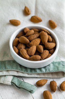 Hafif kumaş peçetede bademlerle dolu beyaz seramik kâse, yumuşak ışıklı ahşap masa arka planı.