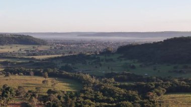 İnsansız hava aracı görüntüleri kırsal tarım arazileri ve yeşil tarlalar üzerinden tepeler ve kırsal bölgelerle çevrili uzak küçük bir kasabaya doğru uçuyor..