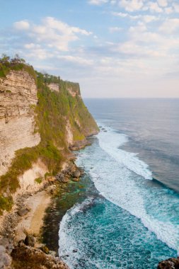 Endonezya, Bali 'de mavi denizli Uluwatu uçurumunun manzarası. Kayalık kayalıklar ve deniz dalgaları, üst manzara. Gün batımında deniz manzarası.