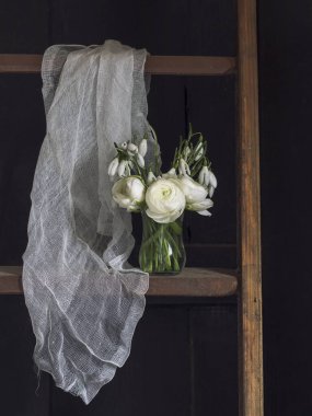 Kar damlaları ve beyaz ranunculuslar koyu arkaplanda cam bir vazoda ve beyaz bir perdenin yanında ahşap bir raf.