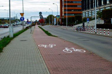19 Ekim 2025, Varşova, Polonya: Arka planda modern ticari binaların bulunduğu işlek bir şehir yoluna paralel uzanan özel bisiklet yolu (kırmızı asfalt) ve yaya kaldırımı (gri asfalt).