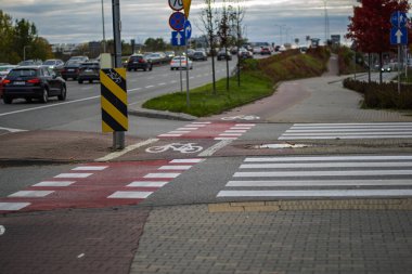 19 Ekim 2025, Varşova, Polonya: Belirli bir yaya geçidi ve yoğun bir şehir caddesinde kırmızı asfalt bir bisiklet geçidi, arabalar ve trafik işaretleri bulutlu bir gökyüzünün altında görülebiliyor.