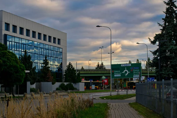 19 Ekim 2025, Varşova, Polonya: Büyük bir otoyol üst geçidinin yanındaki modern cam ve beton ofis binası ve Polonya şehirleri için bulutlu bir gökyüzü altında geniş bir yön tabelası