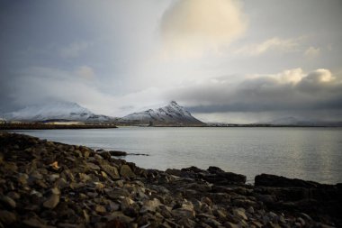 Deniz kenarındaki karla kaplı dağlar dramatik bulutlu gökyüzü altında