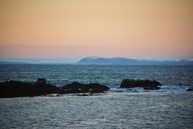 Sunset Coastal View with Distant Snowy Mountains and Rocky Shore