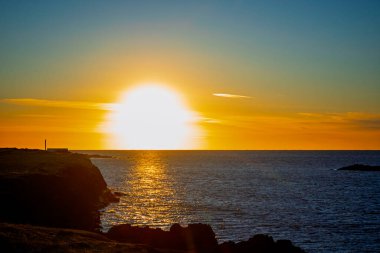 Okyanus ufkunun üzerinde parlak altın gün batımı. Karanlık sahil uçurumları silueti ve parıldayan su yansımaları.