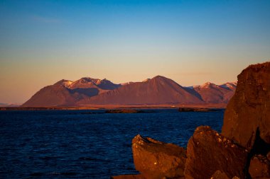 Gün batımında kayalık kıyı şeridi ve derin mavi deniz suyuyla kuzeydeki manzaralı sıradağlar.