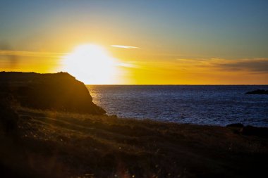 Okyanus ufkunun üzerinde dramatik bir günbatımı. Karanlık siluetli sahil uçurumu ve altın su yansımaları.
