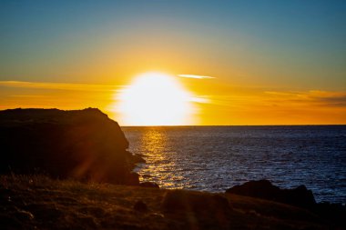 Okyanus ufkunun üzerinde parlak altın gün batımı koyu kayalık kıyı şeridi silueti ve suyun üzerinde güneş yansıması