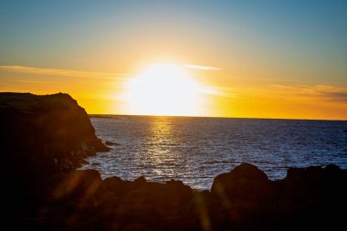 Engebeli kayalık sahillerin üzerinde yoğun altın gün batımı parlak güneş ışığı ve parıldayan okyanus yansımalarıyla