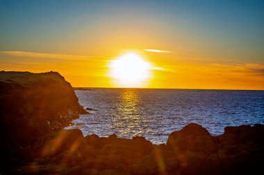Kayalık kıyı şeridi üzerinde dramatik bir gün batımı. Güneş deniz suyuna yansıyor. Canlı altın saat okyanus manzarası.