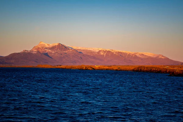 Karla kaplı sıradağlar altın saatinde derin mavi denize tepeden bakıyor, gün batımında manzaralı İskandinav manzarası