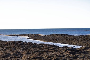 Karanlık volkanik kaya oluşumları ve açık gökyüzünün altında derin mavi deniz suyuyla engebeli kıyı manzarası.