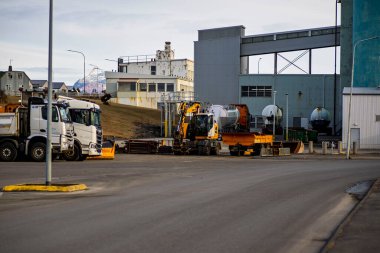 Ağır yük kamyonları ve sarı kazıcı açık bir gökyüzünün altındaki fabrika binalarının yakınındaki bir sanayi bölgesinde park halindeler.