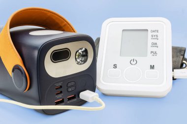 Turuncu saplı siyah portatif bir güç istasyonu, beyaz dijital tansiyon monitörüne bağlanır. Bu da açık mavi arka planda acil tıbbi güç kaynağı kurulumunu gösterir..