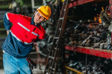 Güvenlik teçhizatındaki bir erkek teknisyen uzun bir fiziksel çalışma gününden sonra yorgunluk ve kas ağrısı belirtileri gösteriyor. Endüstriyel ve mekanik çalışma ortamlarının zorlu doğasını vurguluyor..