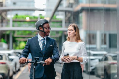 İki iş arkadaşı elektrikli bir scooterla şehir boyunca gidip geliyor, parlak cam binalar arasında ofise doğru gidiyor ve modern, çevre dostu bir iş kültürü sergiliyor..