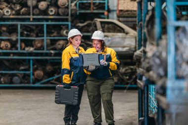 Güvenlik teçhizatı denetlemesi yapan iki kadın teknisyen, engebeli dizüstü bilgisayar kullanarak fabrikada kullanılmış otomobil parçalarını değerlendiriyor. Profesyonel otomotiv ortamında teknik uzmanlık ve takım çalışması gösteriliyor