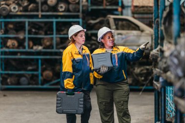 Güvenlik teçhizatı denetlemesi yapan iki kadın teknisyen, engebeli dizüstü bilgisayar kullanarak fabrikada kullanılmış otomobil parçalarını değerlendiriyor. Profesyonel otomotiv ortamında teknik uzmanlık ve takım çalışması gösteriliyor