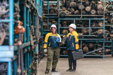 Güvenlik teçhizatı denetlemesi yapan iki kadın teknisyen, engebeli dizüstü bilgisayar kullanarak fabrikada kullanılmış otomobil parçalarını değerlendiriyor. Profesyonel otomotiv ortamında teknik uzmanlık ve takım çalışması gösteriliyor