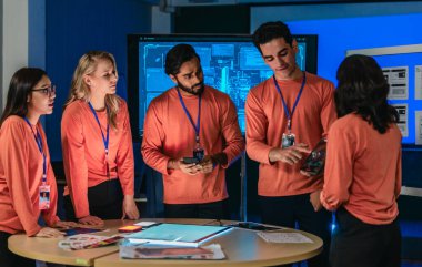 Eşleşen turuncu tişörtlü ürün ekibi masa etrafında işbirliği yapar, kablolar, renk örnekleri, analitik panelli mobil uygulama prototipleri gözden geçirir. UX UI sürat planlama oturumu yakalanıyor