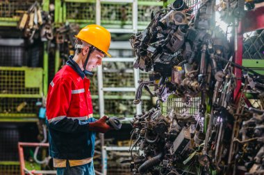 Erkek teknisyen, depo raflarındaki otomotiv parçalarının envanterini incelemek ve kontrol etmek için barkod tarayıcısı ve dijital tablet kullanır.