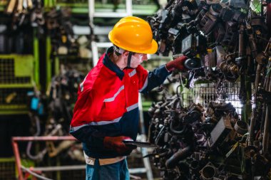 Erkek teknisyen, depo raflarındaki otomotiv parçalarının envanterini incelemek ve kontrol etmek için barkod tarayıcısı ve dijital tablet kullanır.