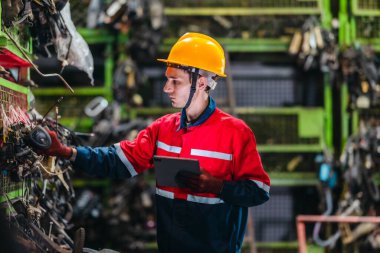 Erkek teknisyen, depo raflarındaki otomotiv parçalarının envanterini incelemek ve kontrol etmek için barkod tarayıcısı ve dijital tablet kullanır.