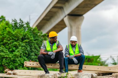 Güvenlik yelekleri ve miğferli iki demiryolu işçisi beton kirişlere ara vererek yorulduklarını gösterirken, aynı zamanda birbirlerini teskin ediyorlar..