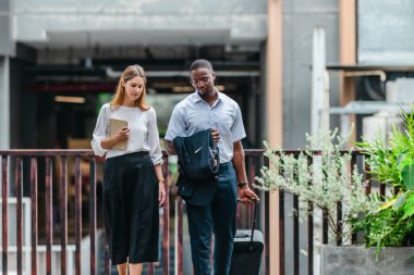 İki profesyonel modern şehir şeridinde yürüyor ve konuşuyor. Şirket seyahatleri, iş seyahatleri ve çevik iş hareketliliğinin sinyallerini veren bir bavul çekiyorlar..