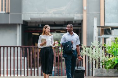 İki profesyonel modern şehir şeridinde yürüyor ve konuşuyor. Şirket seyahatleri, iş seyahatleri ve çevik iş hareketliliğinin sinyallerini veren bir bavul çekiyorlar..