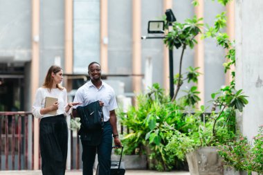 İki profesyonel modern şehir şeridinde yürüyor ve konuşuyor. Şirket seyahatleri, iş seyahatleri ve çevik iş hareketliliğinin sinyallerini veren bir bavul çekiyorlar..