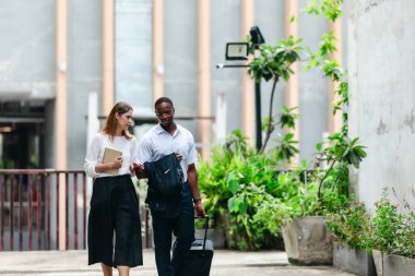İki profesyonel modern şehir şeridinde yürüyor ve konuşuyor. Şirket seyahatleri, iş seyahatleri ve çevik iş hareketliliğinin sinyallerini veren bir bavul çekiyorlar..