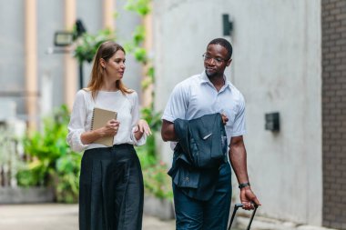 İki profesyonel modern şehir şeridinde yürüyor ve konuşuyor. Şirket seyahatleri, iş seyahatleri ve çevik iş hareketliliğinin sinyallerini veren bir bavul çekiyorlar..