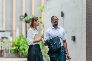 İki profesyonel modern şehir şeridinde yürüyor ve konuşuyor. Şirket seyahatleri, iş seyahatleri ve çevik iş hareketliliğinin sinyallerini veren bir bavul çekiyorlar..