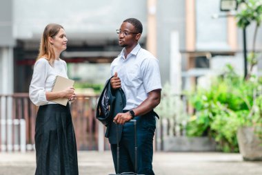 İki profesyonel modern şehir şeridinde yürüyor ve konuşuyor. Şirket seyahatleri, iş seyahatleri ve çevik iş hareketliliğinin sinyallerini veren bir bavul çekiyorlar..