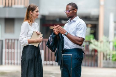 İki profesyonel modern şehir şeridinde yürüyor ve konuşuyor. Şirket seyahatleri, iş seyahatleri ve çevik iş hareketliliğinin sinyallerini veren bir bavul çekiyorlar..