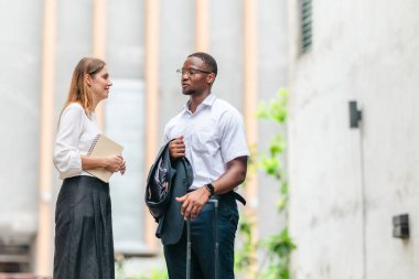 İki profesyonel modern şehir şeridinde yürüyor ve konuşuyor. Şirket seyahatleri, iş seyahatleri ve çevik iş hareketliliğinin sinyallerini veren bir bavul çekiyorlar..
