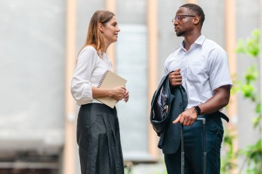 İki profesyonel modern şehir şeridinde yürüyor ve konuşuyor. Şirket seyahatleri, iş seyahatleri ve çevik iş hareketliliğinin sinyallerini veren bir bavul çekiyorlar..