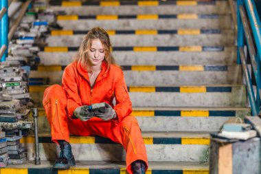 Turuncu kapaklı bir kadın teknisyen tehlikeli merdivenlerde oturur ve yedek eldiveni olan araç kontrol modüllerini parçalar deposunda inceler.