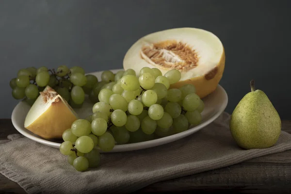 Piring putih dengan melon dan anggur di sebelah pir. Klasik masih hidup. Cahaya lembut. Stok Gambar