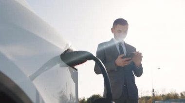 Kafkasyalı işadamı akıllı telefon kullanıyor ve elektrikli araçlara arabadaki aküyü şarj etmek için güç kaynağı bekliyor. Bir elektrikli arabayı şarj istasyonundan şarj etmek