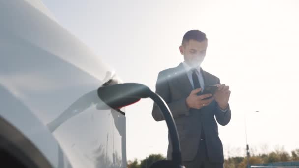 O homem de negócios caucasiano que usa o telefone inteligente e a fonte de alimentação de espera conecta-se aos veículos elétricos para carregar a bateria no carro. Plug carregando um carro elétrico da estação de carregamento — Vídeo de Stock