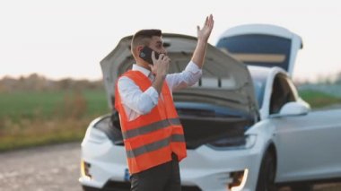 Elektrikli arabası bozulmuş bir adam yardım istiyor. Genç adam telefonda konuşuyor. Araba yardım servisini arıyor. Bozuk arabanın yanında..