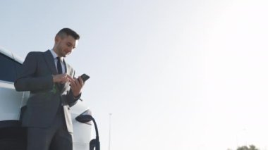 Kafkas işadamı akıllı telefon kullanıyor ve şarj olan beyaz lüks elektrikli arabaya yaslanıyor. Elektrikli arabayı şarj eden fiş.