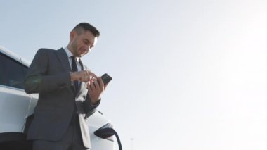 Akıllı telefon kullanan ve şarj olan beyaz elektrikli arabaya yaslanan bir iş adamı. Lüks elektrikli araba şarj oluyor. Elektrikli arabayı şarj eden fiş.