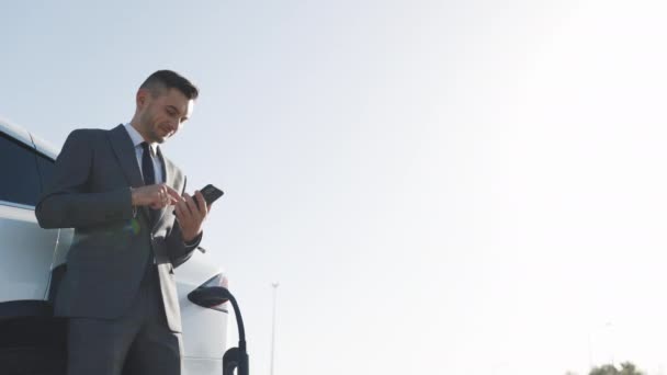 Uomo d'affari caucasico utilizzando smart phone e si appoggia su bianco auto elettrica di lusso che si sta caricando. Spina di ricarica di un'auto elettrica — Video Stock