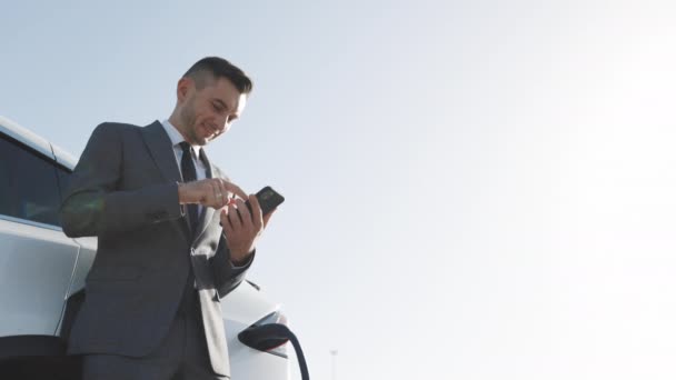 Επιχειρηματίας χρησιμοποιώντας smartphone και γέρνει σε λευκό πολυτελές ηλεκτρικό αυτοκίνητο που φορτίζει. Πολυτελές ηλεκτρικό αυτοκίνητο επαναφόρτιση. Φορτίστε ένα ηλεκτρικό αυτοκίνητο — Αρχείο Βίντεο