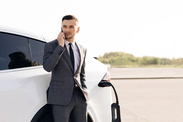 Porträtt av stilig glad 30-årig affärsman med snyggt skägg som har mobil konversation medan egen lyxig bil laddning batteri på speciellt utrustade laddstation — Stockfoto