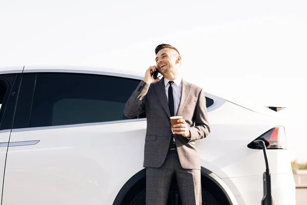 Empresário está falando em seu telefone inteligente, enquanto seu carro elétrico está carregando no fundo. Vista lateral do carro de luxo branco. Ambientalmente consciente masculino carregamento veículo elétrico — Fotografia de Stock